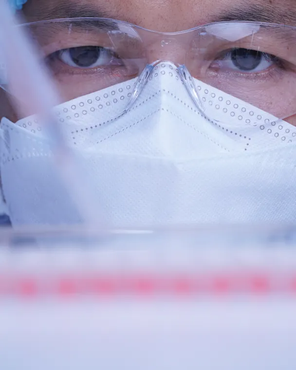 a scientist in a mask, hairnet and safety glasses pipetting into a tray of tubes