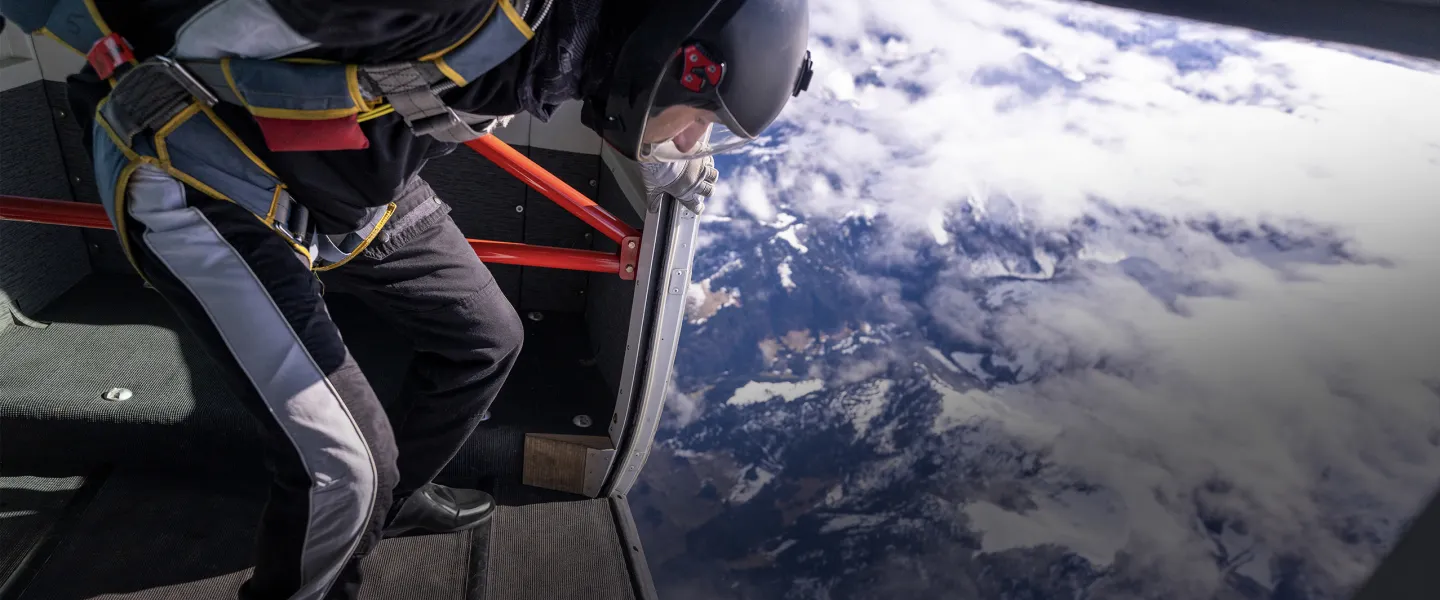 a skydiver in full safety gear standing at a plane door overlooking clouds in the sky