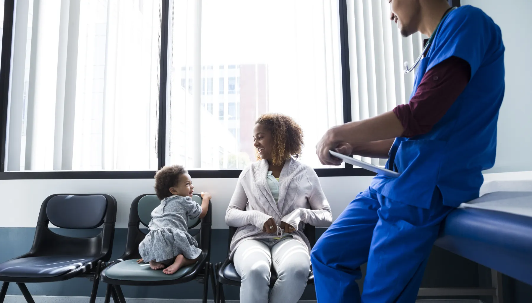 Small child looks out the window of a hospital room while a caregiver and healthcare worker look on