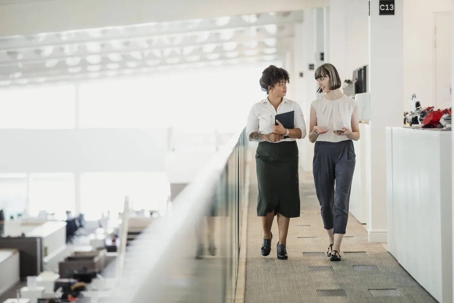 Two female work colleagues talk as they walk down an office hallway